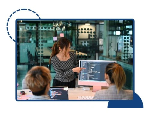 A team collaborating in a modern office where a developer is pointing at code displayed on a monitor while explaining a project to colleagues — showcasing Custom PHP Web Development Services by Digital Dividend.